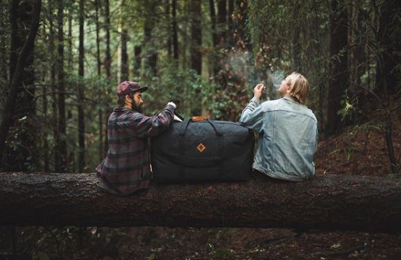 two travelers enjoying a smoke on a log with a reverly bag between them